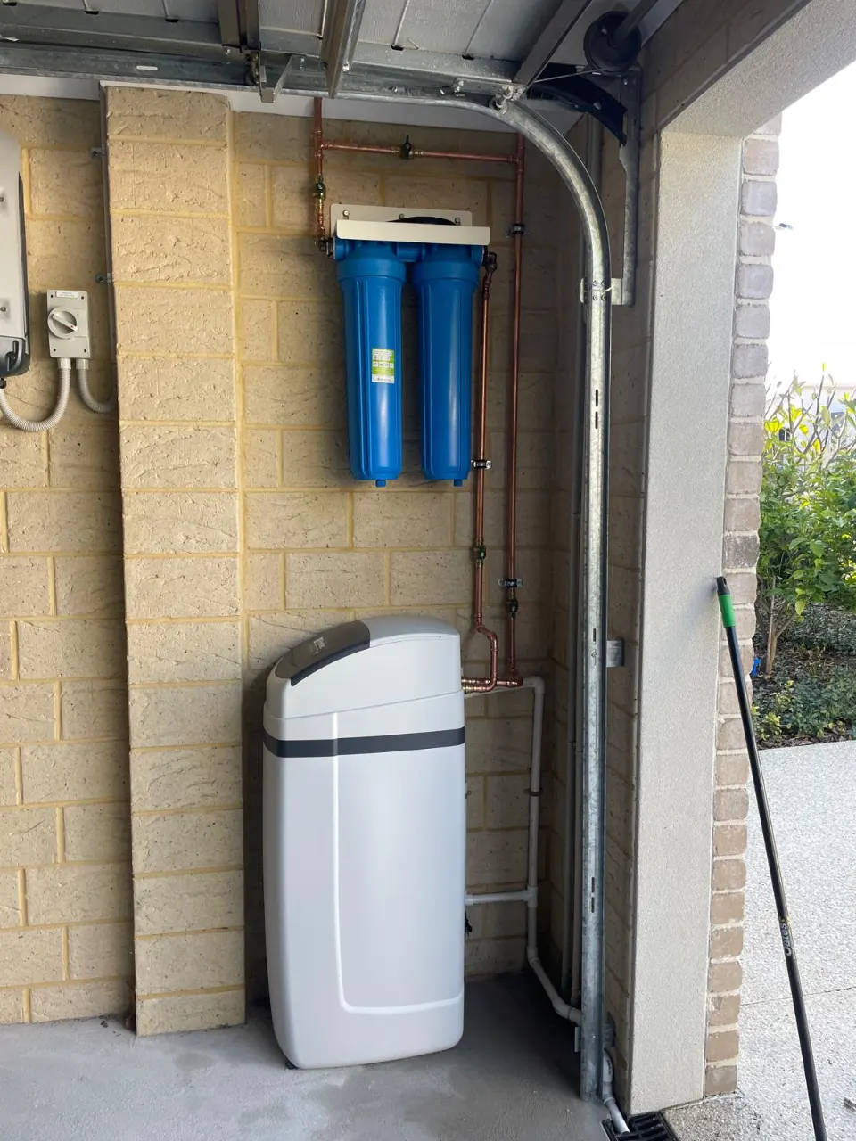 Two blue whole-house water filter housings above a water softener in a garage, with copper pipework and conduit