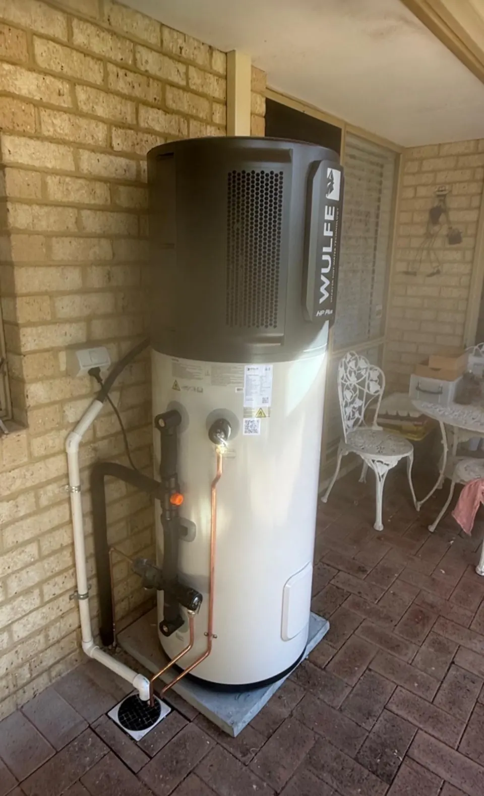 Wulfe heat pump hot water system on a paved patio against a brick wall, with insulated pipework and a drain