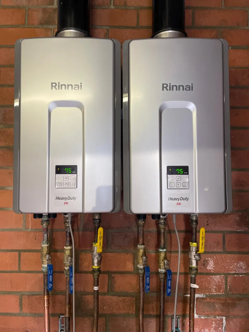 Two Rinnai HeavyDuty continuous flow gas hot water units on a brick wall with gas isolation valves and copper pipework below