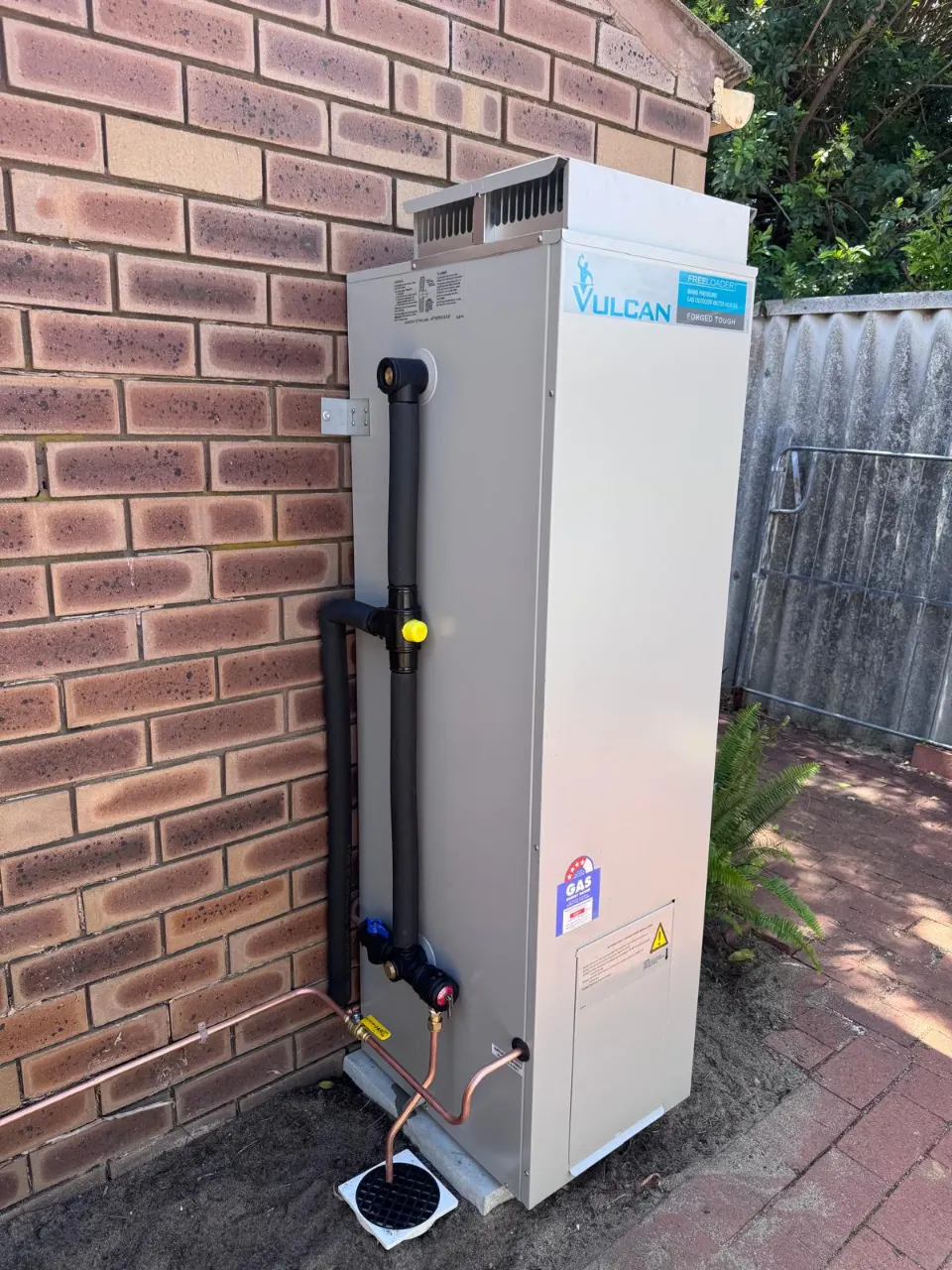 Vulcan gas storage hot water system on a red brick wall beside a paved path, with insulated pipework and a copper pressure-relief line running to a drain